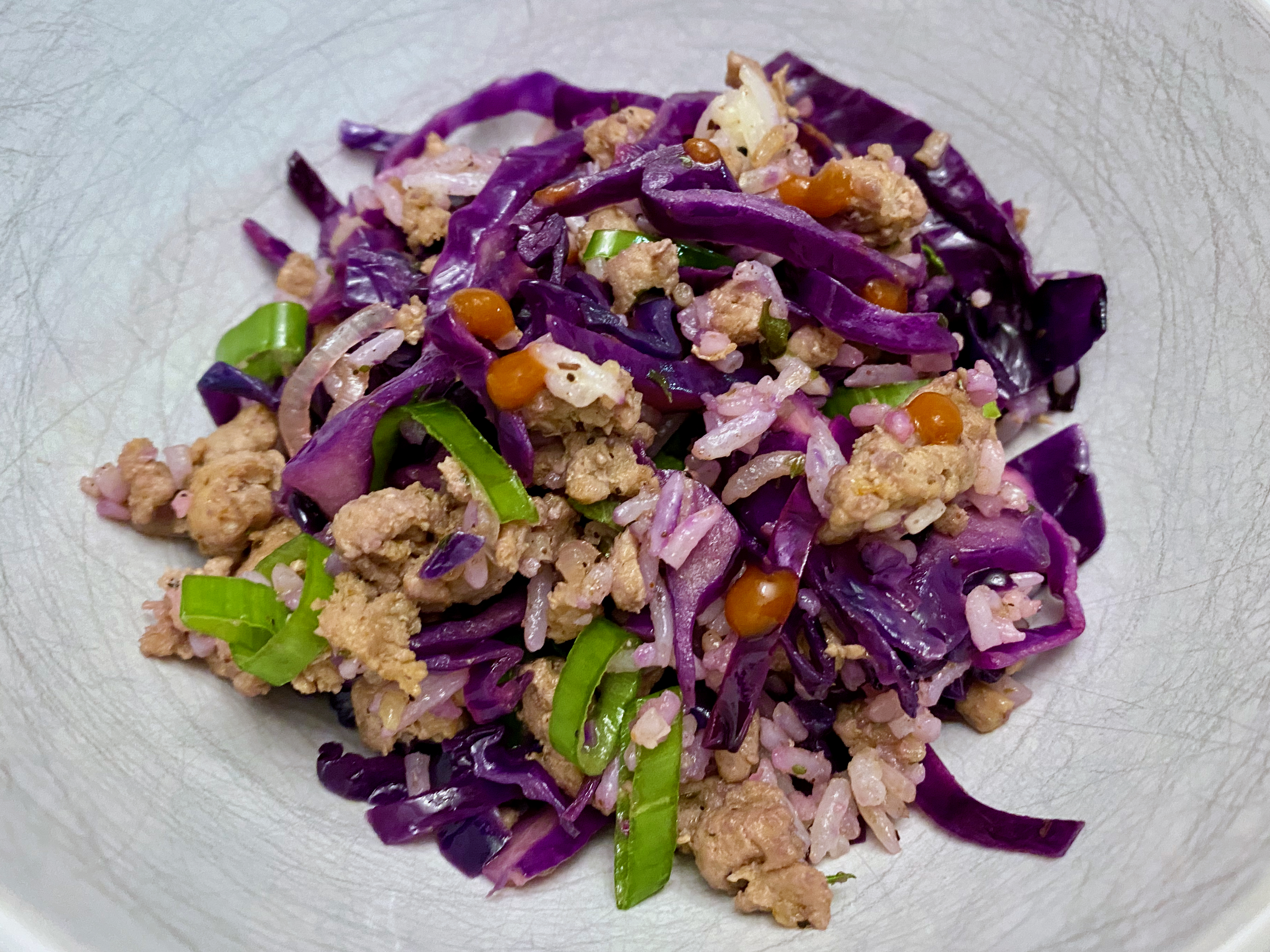 Purple cabbage, green onion, browned meat, rice, and red sriracha dots in a white bowl.