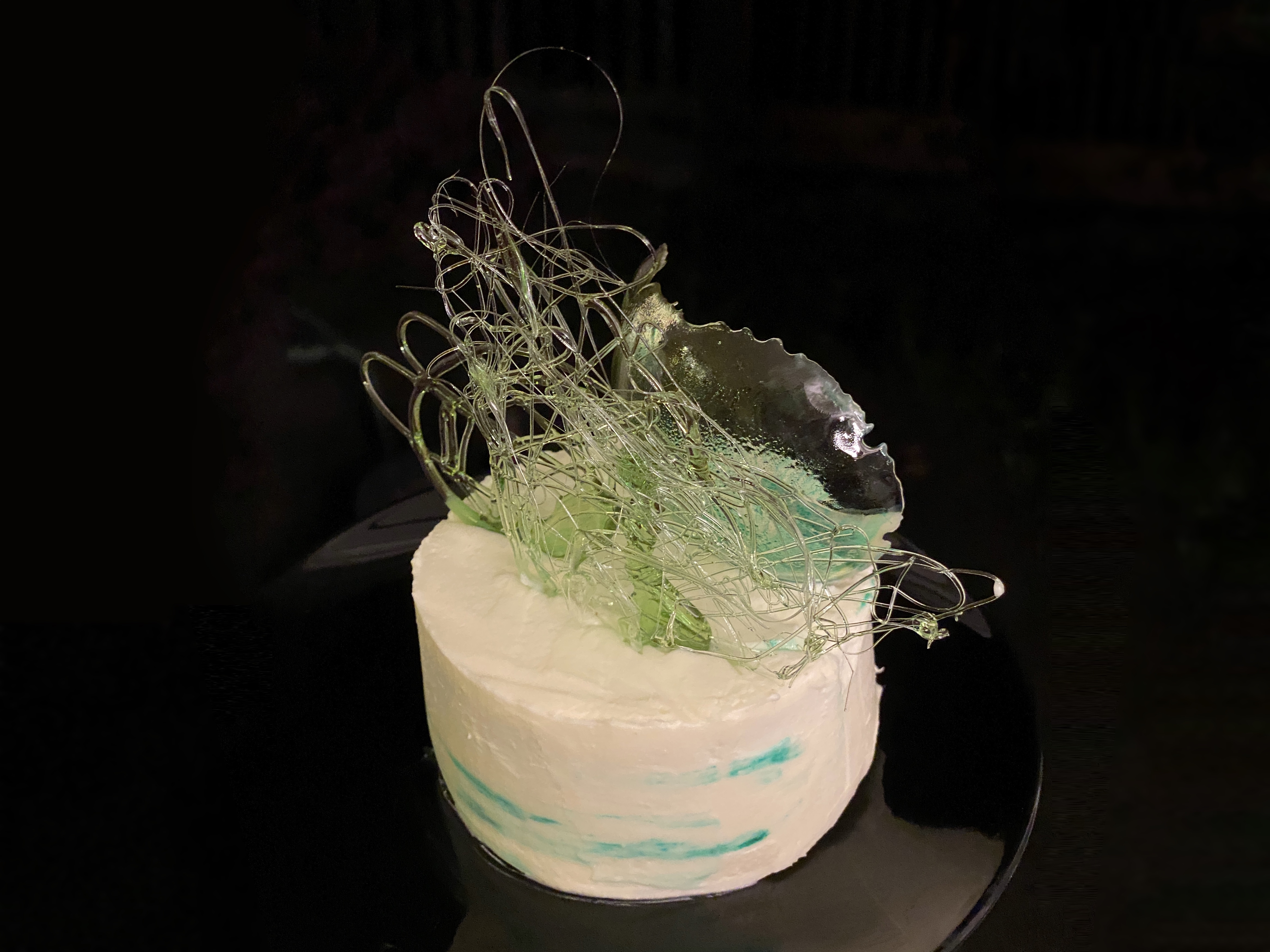 Tall, round cake, with teal on white watercolor style frosting, topped with green and teal abstract sugar work.